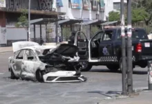 A Mexican military convoy with armored vehicles and armed soldiers parked on a highway with smoke in the distance.