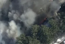 An aerial view of a large industrial building with a partially collapsed roof and black smoke rising into the air.
