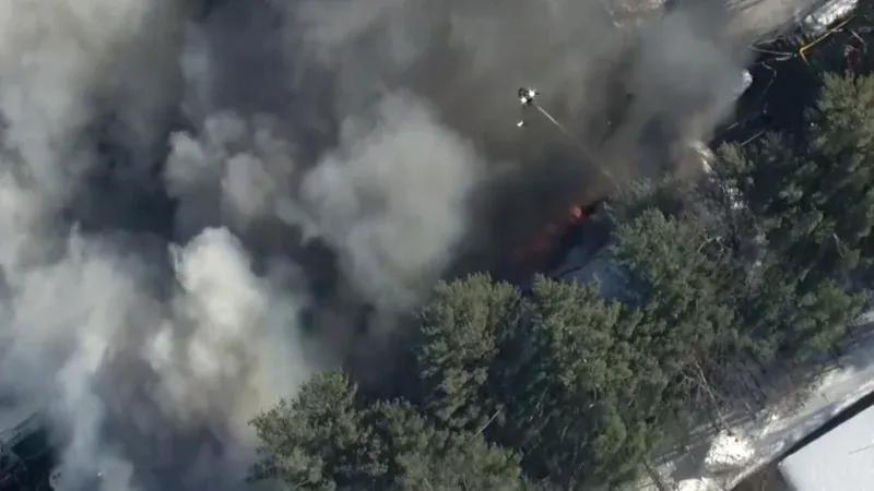 An aerial view of a large industrial building with a partially collapsed roof and black smoke rising into the air.